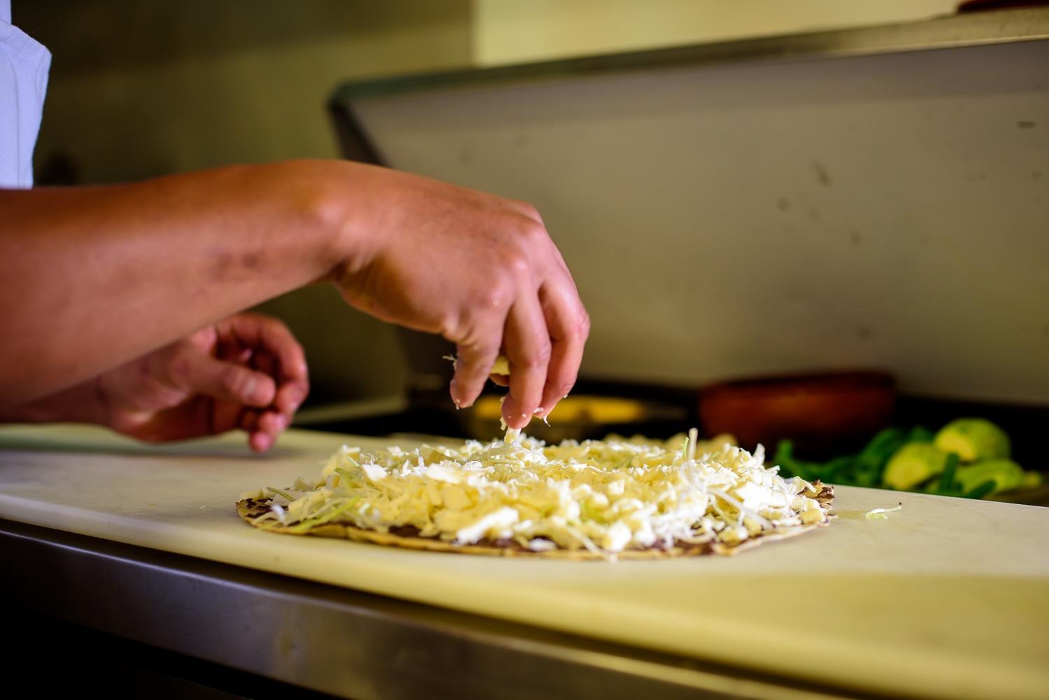 Manos esparciendo quesillo desmenuzado sobre una memela con frijoles, en una cocina oaxaqueña